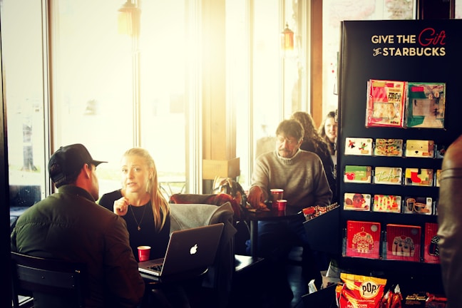 Several people are seated in a coffee shop with large windows allowing in natural light. A man and woman are engaged in conversation at a table, where a laptop is open in front of the woman. The man is wearing a hat and a jacket, while the woman has long blonde hair and is holding a small red cup. A display stand offers Starbucks gift cards. In the background, other patrons are sitting at tables enjoying their beverages.