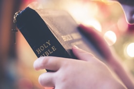 A person is holding a black leather-bound book with the title 'HOLY BIBLE' embossed in gold letters. A delicate gold cross charm is attached to the book, and the background features soft, out-of-focus lights that create a warm and serene atmosphere.