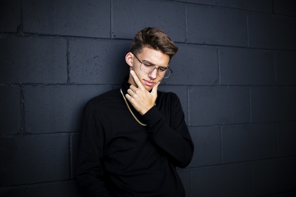 man leaning on wall while looking down wearing eyeglasses and necklace with right hand on chin
