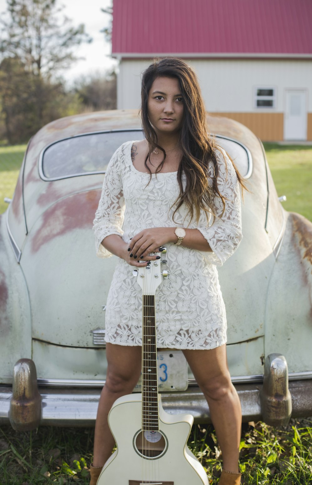 A woman in a dress holding a guitar in front of an older white car.
