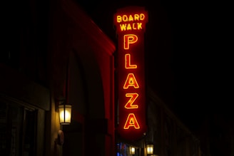 A neon sign with the words 'Board Walk Plaza' in bright red letters is illuminated against a dark background. The sign is vertically oriented, and several lantern-style wall lights near the building provide additional light.