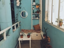A cozy indoor nook with turquoise walls is adorned with potted plants and a small seating area. The corner features a white bench with a patterned cushion and several small potted plants arranged around it. A circular mirror hangs on the wall above a series of shelves holding more plants and decorative items. Large windows on the right let in natural light, illuminating the space.
