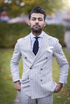 A man stands confidently outdoors wearing a light gray pinstripe suit with a white dress shirt and a dark tie. The suit is double-breasted and includes a pocket square. His hands are in his pockets, and he has a neatly groomed beard and hair. The background has blurred greenery and hints of autumn leaves.