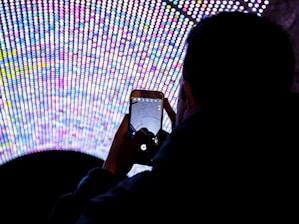 A person is holding a smartphone, capturing a photo of a large, vibrant LED display composed of numerous small, colorful lights arranged in a semi-circular pattern. The scene is viewed from behind the person, highlighting the bright, multicolored lights against the dark surroundings.