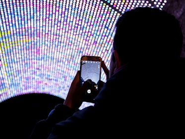 A person is holding a smartphone, capturing a photo of a large, vibrant LED display composed of numerous small, colorful lights arranged in a semi-circular pattern. The scene is viewed from behind the person, highlighting the bright, multicolored lights against the dark surroundings.
