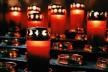 A group of red candles with golden lids are arranged in a dimly lit setting, casting a warm glow around them. The intricate designs on the lids allow light to escape in distinctive patterns, creating an atmosphere of tranquility and contemplation.