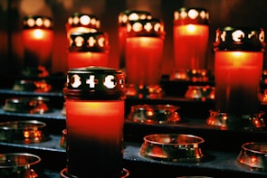A group of red candles with golden lids are arranged in a dimly lit setting, casting a warm glow around them. The intricate designs on the lids allow light to escape in distinctive patterns, creating an atmosphere of tranquility and contemplation.