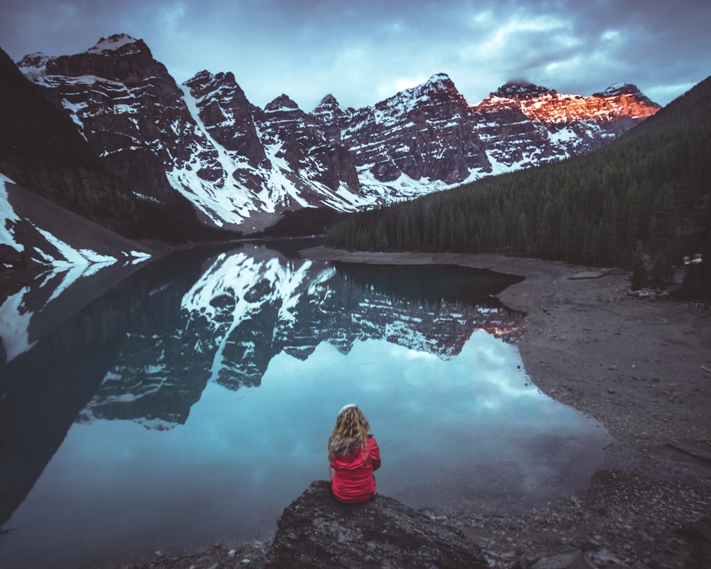 Frau sitzt auf Felsen vor ruhigem Gewässer und Blick auf die Berge
