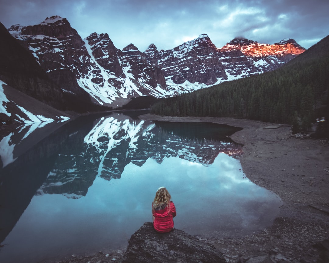 woman sitting on rock in front calm body of water and mountains view, Mornings at Moraine Lake - photo by @kalenemsley a