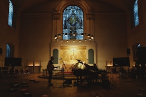 A dimly lit church interior features a grand piano and musician setup at the altar. Stained glass windows illuminate the room with a subtle glow. Microphones and other musical equipment are placed around, suggesting a performance or recording. The atmosphere is quiet and reverent, as illuminated candles line the space, enhancing the sacred ambiance.