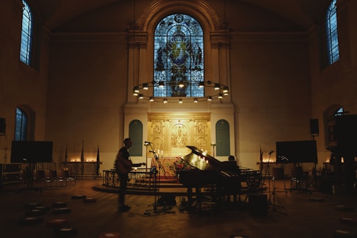 A dimly lit church interior features a grand piano and musician setup at the altar. Stained glass windows illuminate the room with a subtle glow. Microphones and other musical equipment are placed around, suggesting a performance or recording. The atmosphere is quiet and reverent, as illuminated candles line the space, enhancing the sacred ambiance.