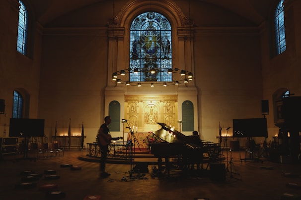 A dimly lit church interior features a grand piano and musician setup at the altar. Stained glass windows illuminate the room with a subtle glow. Microphones and other musical equipment are placed around, suggesting a performance or recording. The atmosphere is quiet and reverent, as illuminated candles line the space, enhancing the sacred ambiance.