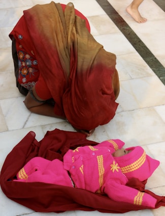 A person wearing a red and brown saree is kneeling on a white tiled floor. In front of them lies a baby dressed in a bright pink outfit with yellow patterns, wrapped in a maroon cloth. The scene conveys a sense of care and attention.