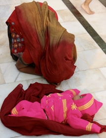 A person wearing a red and brown saree is kneeling on a white tiled floor. In front of them lies a baby dressed in a bright pink outfit with yellow patterns, wrapped in a maroon cloth. The scene conveys a sense of care and attention.