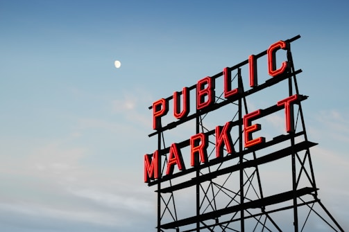 A large metal sign with bright red neon lettering reads 'PUBLIC MARKET' against a clear sky with a visible moon.