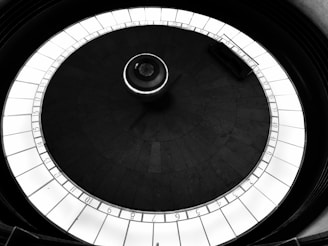 A monochromatic, top-down view of a circular pendulum with a numbered track surrounding it. The central pendulum, suspended above the floor, is enclosed by a bright, segmented ring with numbers from 1 to 44, creating a stark contrast against the darker background.