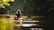 A person is kayaking in a calm river surrounded by dense green foliage. The sunlight filters through the trees, casting a warm glow on the water. The scene is tranquil and serene, with the kayak gliding smoothly across the reflective surface.