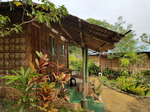 A rustic structure made of woven bamboo and wood with a sloped thatched roof is seen surrounded by lush greenery. The scene includes various tropical plants and shrubs along a narrow stone pathway. The house has a porch area with plastic chairs, and the atmosphere is serene and inviting.
