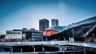 A modern urban landscape featuring a futuristic building with a reflective, undulating facade and a large digital screen displaying 'Grand Central' with a vibrant red background. In the background, there are tall office buildings and a clear blue sky.