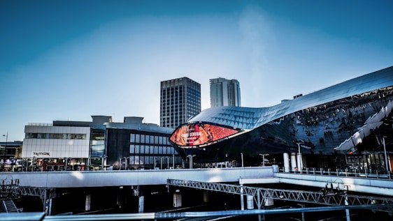 A modern urban landscape featuring a futuristic building with a reflective, undulating facade and a large digital screen displaying 'Grand Central' with a vibrant red background. In the background, there are tall office buildings and a clear blue sky.