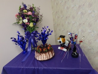 A decorative table setup featuring a chocolate cake adorned with strawberries at the center. To the left, a tall, vibrant bouquet of flowers stands in a vase, including various flowers such as daisies and carnations in shades of purple, pink, yellow, and white. Two smaller blue metallic decorations are placed on either side, matching the deep purple table cover. To the right, a plush toy wearing a graduation cap and holding a diploma sits beside additional colorful metallic decorations.