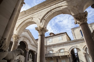 An architectural scene featuring ancient stone structures with ornate arches and columns. The sky is visible in the background, adding a sense of openness and balance. Statues and intricate carvings are present, highlighting historical craftsmanship.