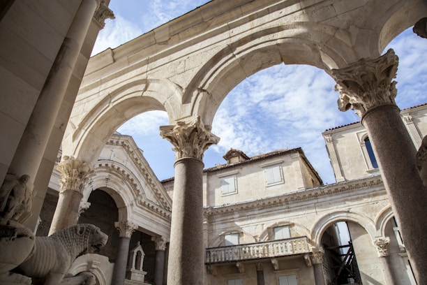An architectural scene featuring ancient stone structures with ornate arches and columns. The sky is visible in the background, adding a sense of openness and balance. Statues and intricate carvings are present, highlighting historical craftsmanship.