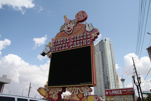 A large, colorful sign featuring a clown with a lollipop and pointing hands stands in front of a backdrop of tall buildings and a clear, partly cloudy sky. The sign reads 'Circus Circus Hotel Casino' and is positioned above a prominent black display screen. Electrical wires and a theme park advertisement are visible in the surroundings.