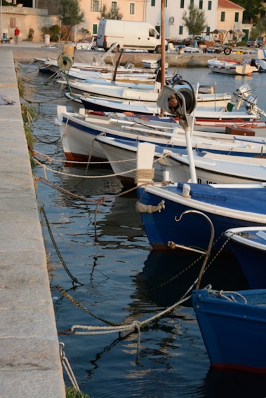 Several small boats are moored along a stone pier in a calm harbor. The boats are primarily white with blue accents, and they are secured with ropes. In the background, there are buildings with light-colored facades and green shutters, along with a parked white van. The water reflects the boats and surroundings in a gentle manner, suggesting a peaceful setting.