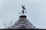 A rooftop with a weather vane shaped like a grasshopper against a cloudy sky. The vane has cardinal directions marked as N, S, E, and W.