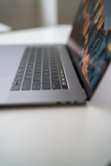 A sleek laptop is partially open on a clean, white surface, with a focus on the keyboard and touchpad. The screen displays a vibrant, digital image with abstract patterns.