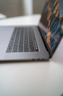 A sleek laptop is partially open on a clean, white surface, with a focus on the keyboard and touchpad. The screen displays a vibrant, digital image with abstract patterns.