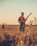 Two individuals are walking through a field of tall, dry grass under a clear sky. They are dressed in outdoor gear, including bright orange vests, and carrying shotguns. The field stretches out under the warm light of late afternoon or early evening, with bare trees visible in the distance.