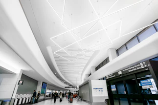 A modern airport terminal features a sleek design with a high ceiling adorned with geometric lighting. People walk with luggage towards the entrance and kiosks, creating a busy yet orderly atmosphere. The signage indicates directions for terminals and parking shuttles.