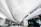 A modern airport terminal features a sleek design with a high ceiling adorned with geometric lighting. People walk with luggage towards the entrance and kiosks, creating a busy yet orderly atmosphere. The signage indicates directions for terminals and parking shuttles.