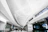 A modern airport terminal features a sleek design with a high ceiling adorned with geometric lighting. People walk with luggage towards the entrance and kiosks, creating a busy yet orderly atmosphere. The signage indicates directions for terminals and parking shuttles.