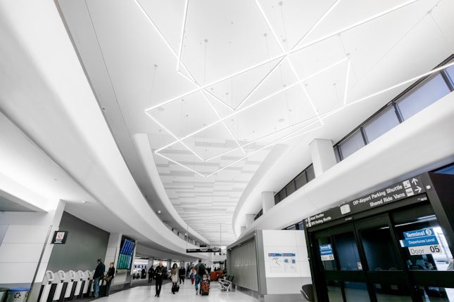 A modern airport terminal features a sleek design with a high ceiling adorned with geometric lighting. People walk with luggage towards the entrance and kiosks, creating a busy yet orderly atmosphere. The signage indicates directions for terminals and parking shuttles.