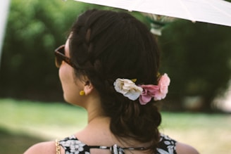 A person with dark braided hair adorned with flower clips, wearing sunglasses and a floral-patterned outfit. They are holding a white umbrella, with a blurred green outdoor background.