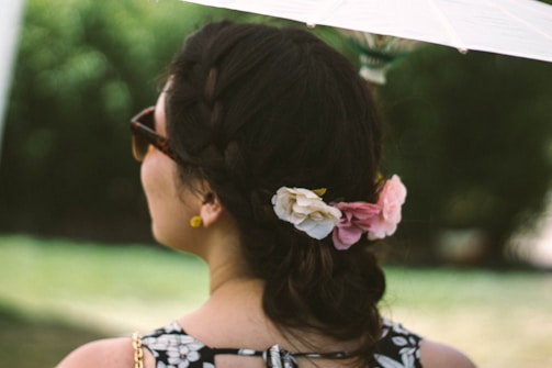 A person with dark braided hair adorned with flower clips, wearing sunglasses and a floral-patterned outfit. They are holding a white umbrella, with a blurred green outdoor background.