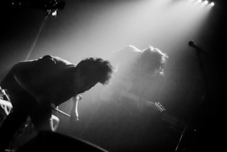 A dramatic black and white image capturing two musicians in a dimly lit setting. One appears to be bending over or crouched, possibly presenting an energetic or intense performance. The other is playing a guitar, partially obscured by hair and shadows. The scene is filled with smoky or misty light, creating a moody atmosphere, with a microphone stand visible on the right.
