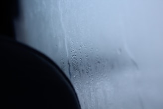 A foggy glass surface with visible condensation and water droplets. The image features a soft gradient from a darker area on the left to a lighter, airy blue on the right, suggesting a cold or humid environment.