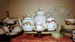 An assortment of vintage porcelain teapots, teacups, and saucers displayed on a shelf. The teapots and cups feature delicate floral patterns, predominantly with roses. The arrangement includes a large serving plate in the background and several stacked floral-patterned plates and bowls. The items are placed on a lace tablecloth, emphasizing a classic, elegant style.