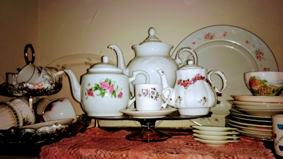 An assortment of vintage porcelain teapots, teacups, and saucers displayed on a shelf. The teapots and cups feature delicate floral patterns, predominantly with roses. The arrangement includes a large serving plate in the background and several stacked floral-patterned plates and bowls. The items are placed on a lace tablecloth, emphasizing a classic, elegant style.