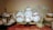 An assortment of vintage porcelain teapots, teacups, and saucers displayed on a shelf. The teapots and cups feature delicate floral patterns, predominantly with roses. The arrangement includes a large serving plate in the background and several stacked floral-patterned plates and bowls. The items are placed on a lace tablecloth, emphasizing a classic, elegant style.
