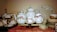 An assortment of vintage porcelain teapots, teacups, and saucers displayed on a shelf. The teapots and cups feature delicate floral patterns, predominantly with roses. The arrangement includes a large serving plate in the background and several stacked floral-patterned plates and bowls. The items are placed on a lace tablecloth, emphasizing a classic, elegant style.