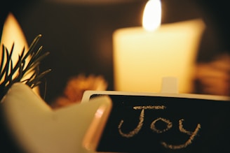 A lit candle is in the background creating a warm and cozy atmosphere. In the foreground, a small blackboard with the word 'Joy' written in chalk is visible. Pine leaves are also present on the side, contributing to a festive or holiday feel.