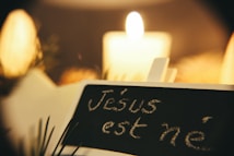 A softly lit scene with a candle glowing warmly in the background. In the foreground, a chalkboard reads 'J&eacute;sus est n&eacute;,' suggesting a festive, possibly Christmas-related theme. Pine needles partially frame the image, hinting at seasonal decor.