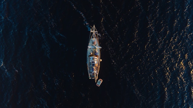 Aerial view of a sailboat with a small attached dinghy floating on dark blue ocean water. The boat is centered in the image, showing the vessel's deck and masts clearly, while the surrounding water reflects some golden sunlight.