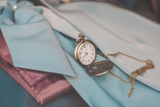 A vintage pocket watch with an ornate design rests on fabric in shades of teal and purple. The watch chain is intricately looped and creates a charming contrast with the surrounding materials.