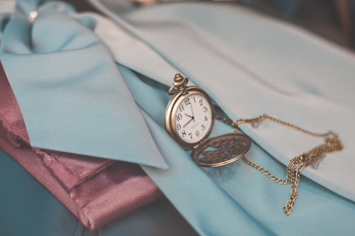 A vintage pocket watch with an ornate design rests on fabric in shades of teal and purple. The watch chain is intricately looped and creates a charming contrast with the surrounding materials.